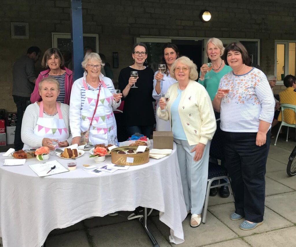 Image shows WI members outside of Tackley Village Hall holding glasses of wine. There is a table in the foreground with slices of cake on plates