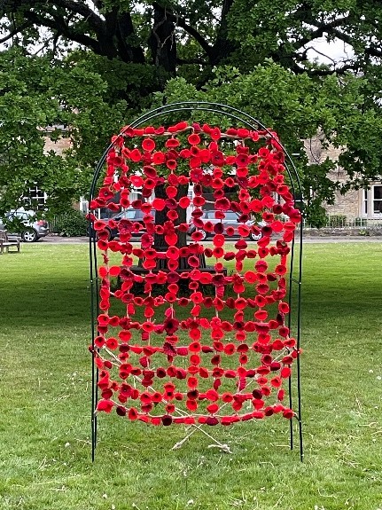 Image shows an oval garden trellis covered in poppies crocheted or knitted by Tackley WI members. The trellis is on the village green with the large oak tree behind